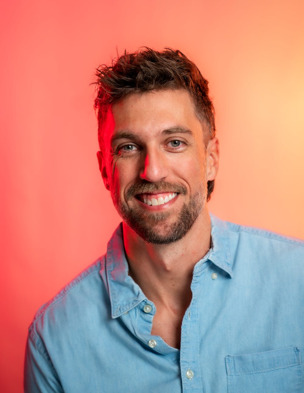 Nathan smiling in a chambray shirt against a warm pink and amber background
