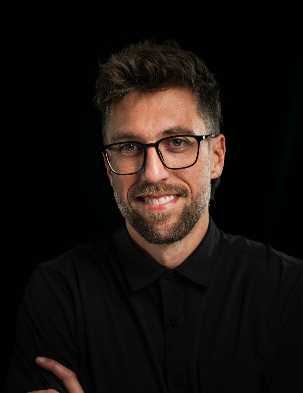 Nathan smiling in glasses and a black button-up shirt, against a black background
